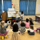 Children sitting on the floor, listening to a teacher read a book